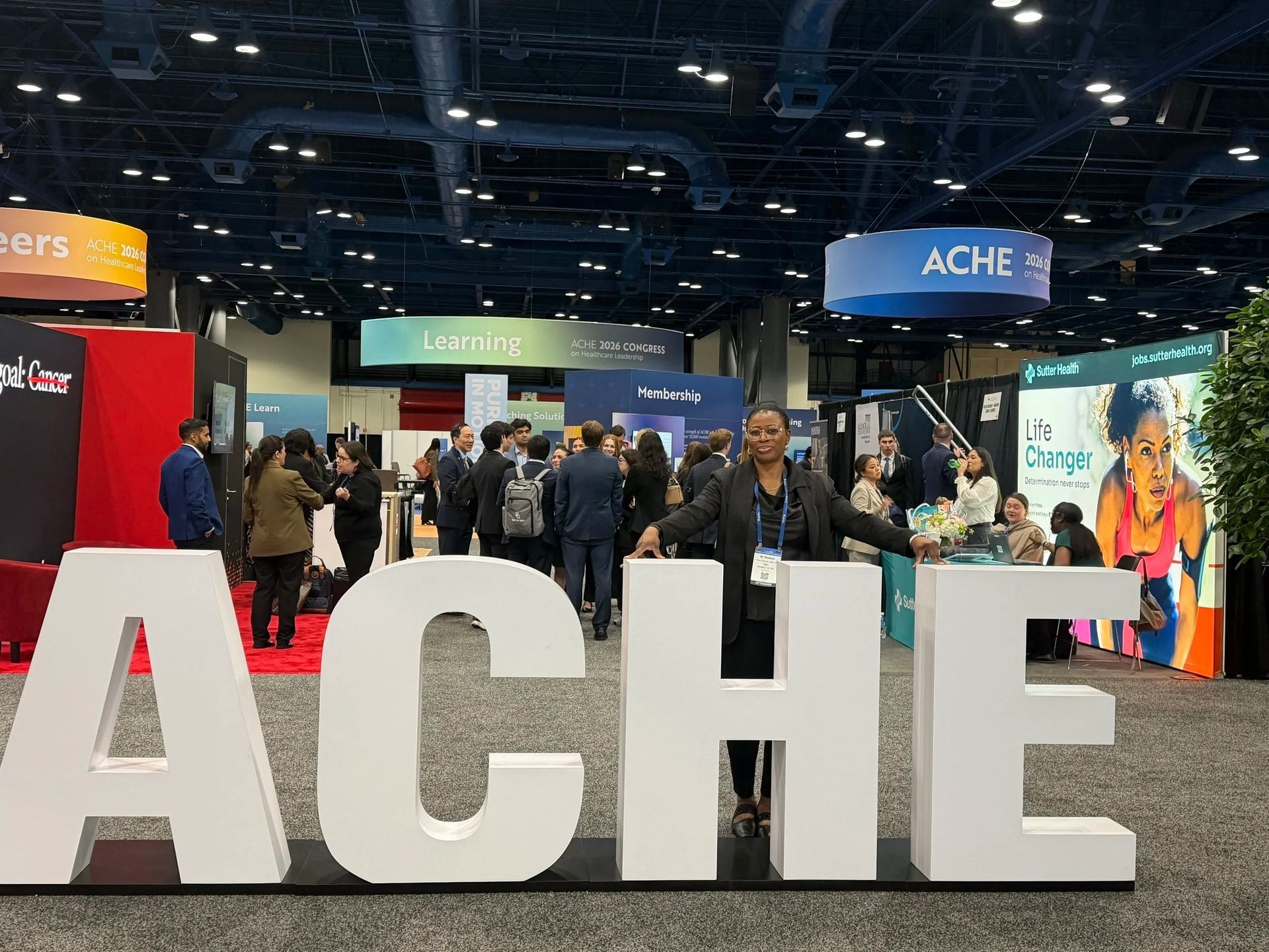 ACHE conference exhibit hall with large white letters and attendees browsing booths