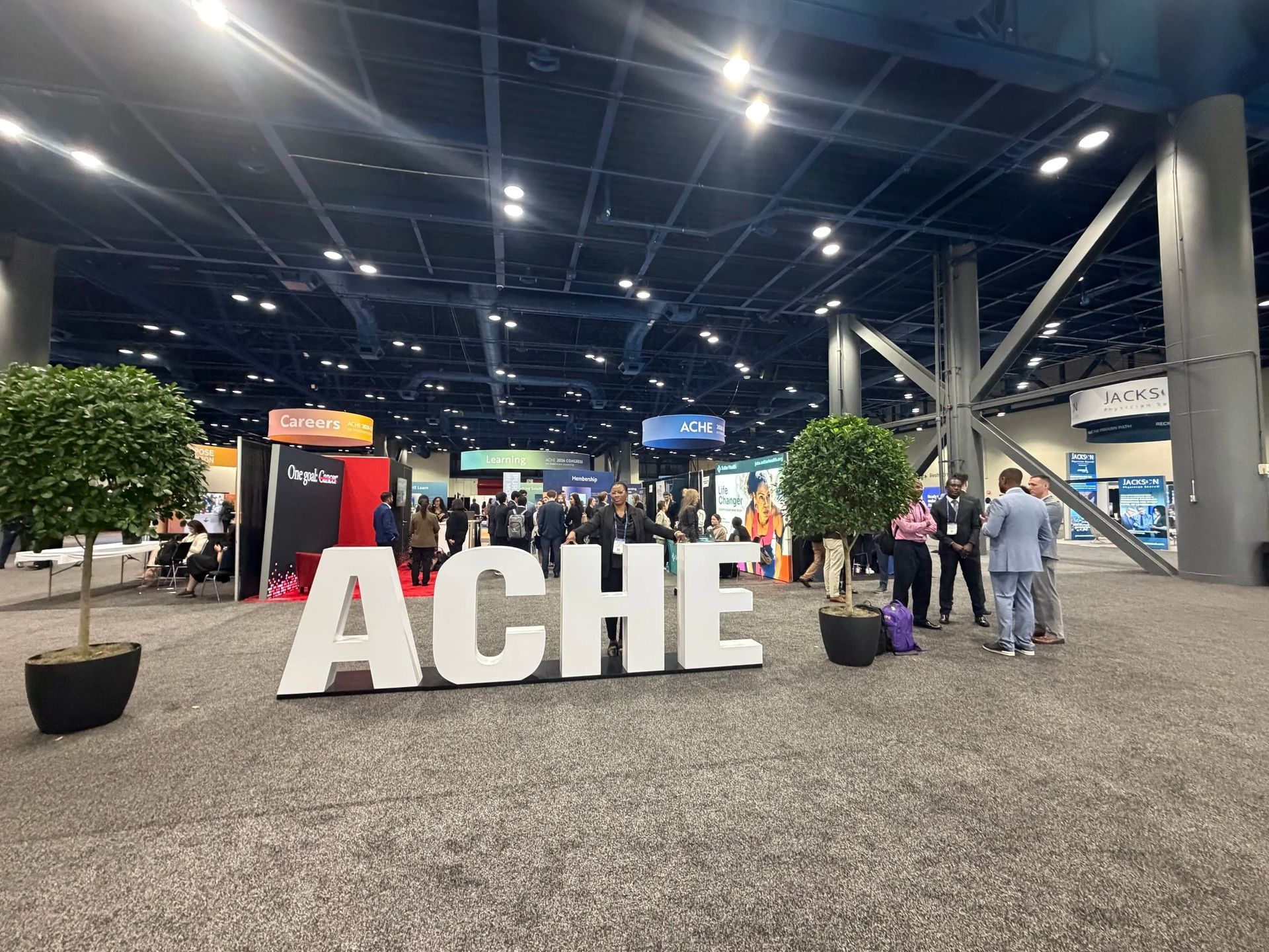 Large “ACHE” letters in a convention hall with people walking and overhead lights.