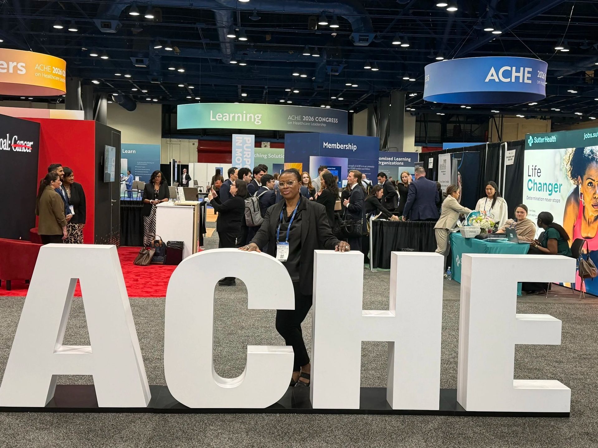 People at a trade show behind large white “ACHE” letters in a busy exhibition hall.
