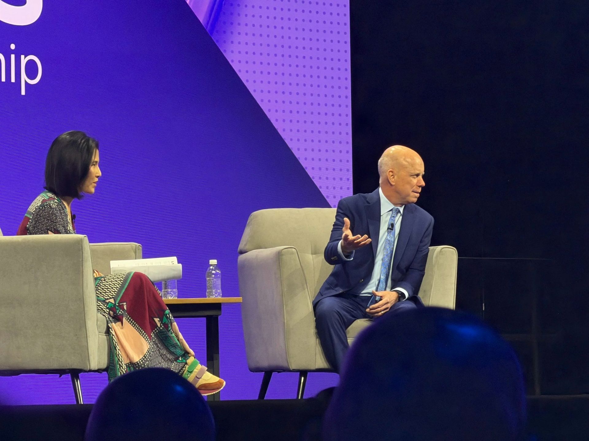 Two speakers on stage in armchairs during a panel discussion, one gesturing while seated under purple lighting