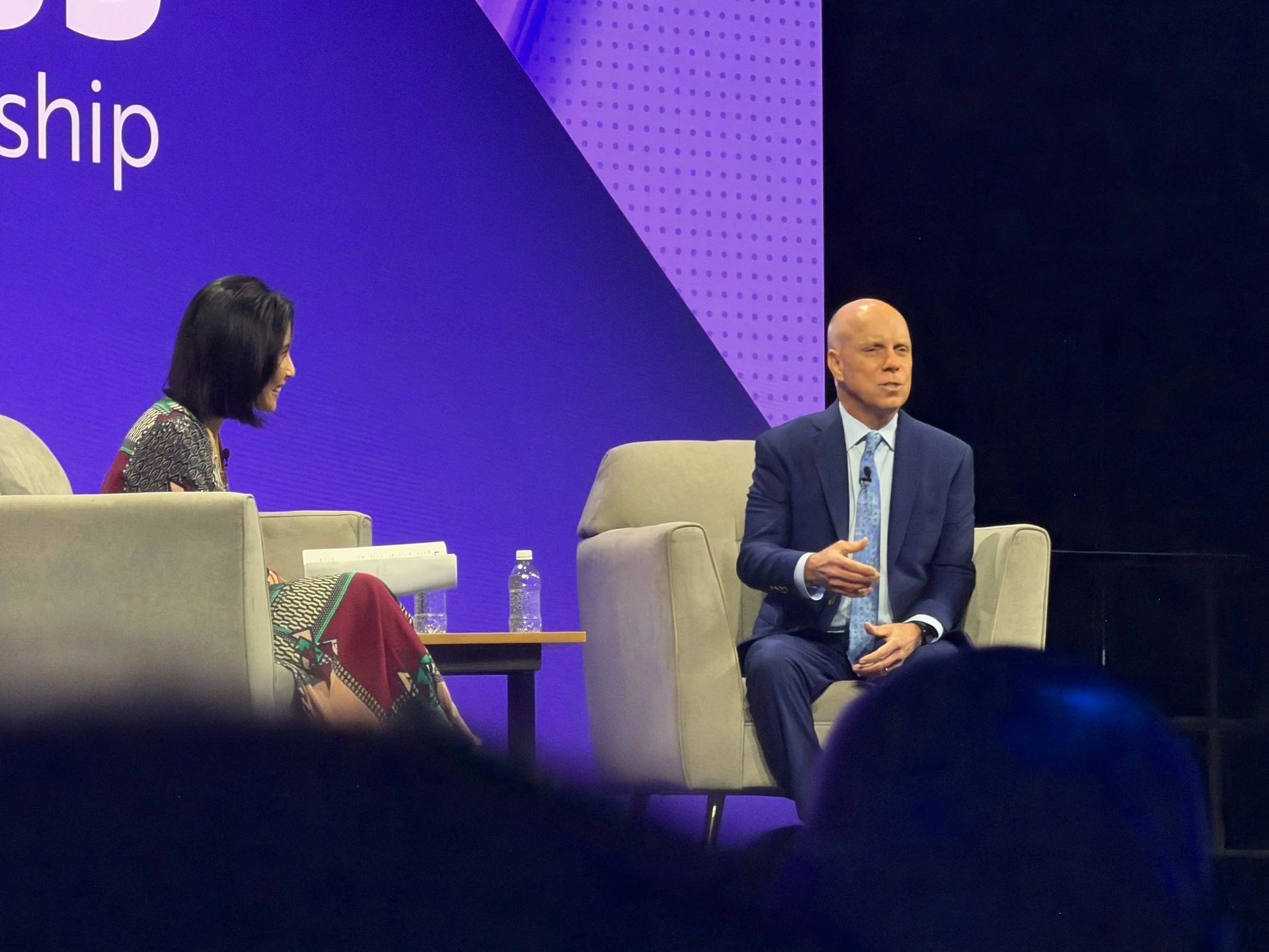 Two people on stage in gray chairs during a panel discussion, with a purple backdrop and a table between them.
