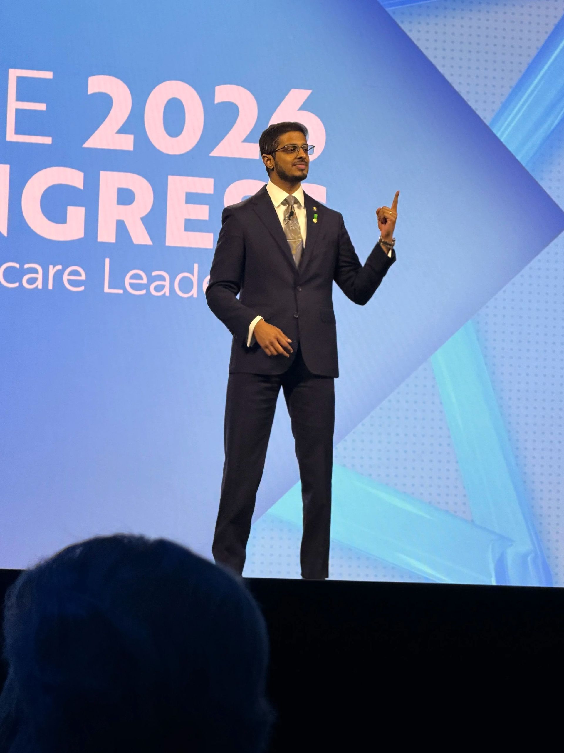 Speaker on stage at a 2025 healthcare leadership conference, standing before a blue backdrop and gesturing with one hand.