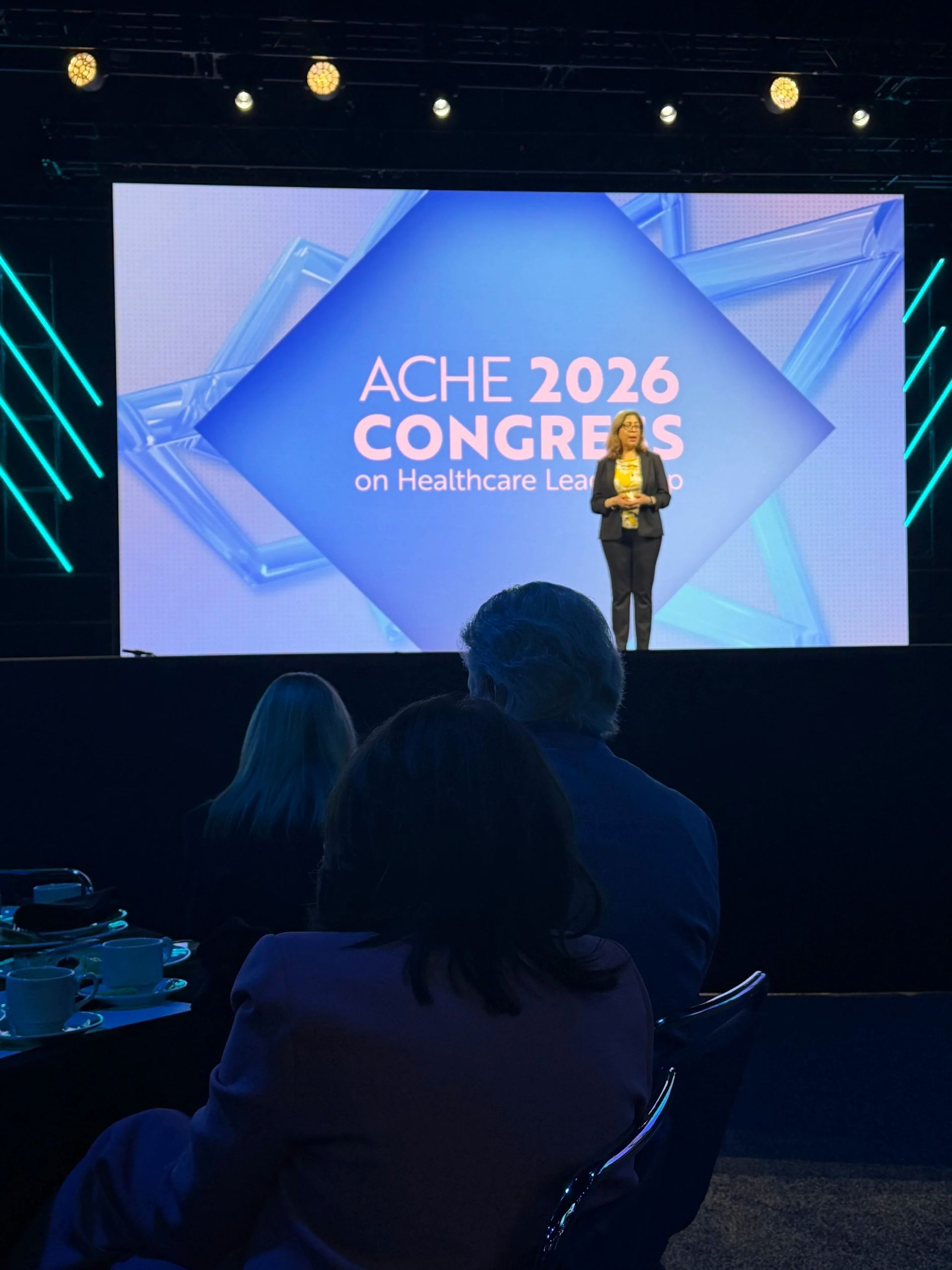 Speaker on stage at ACHE 2026 Congress in front of a blue-lit screen, viewed from the audience.
