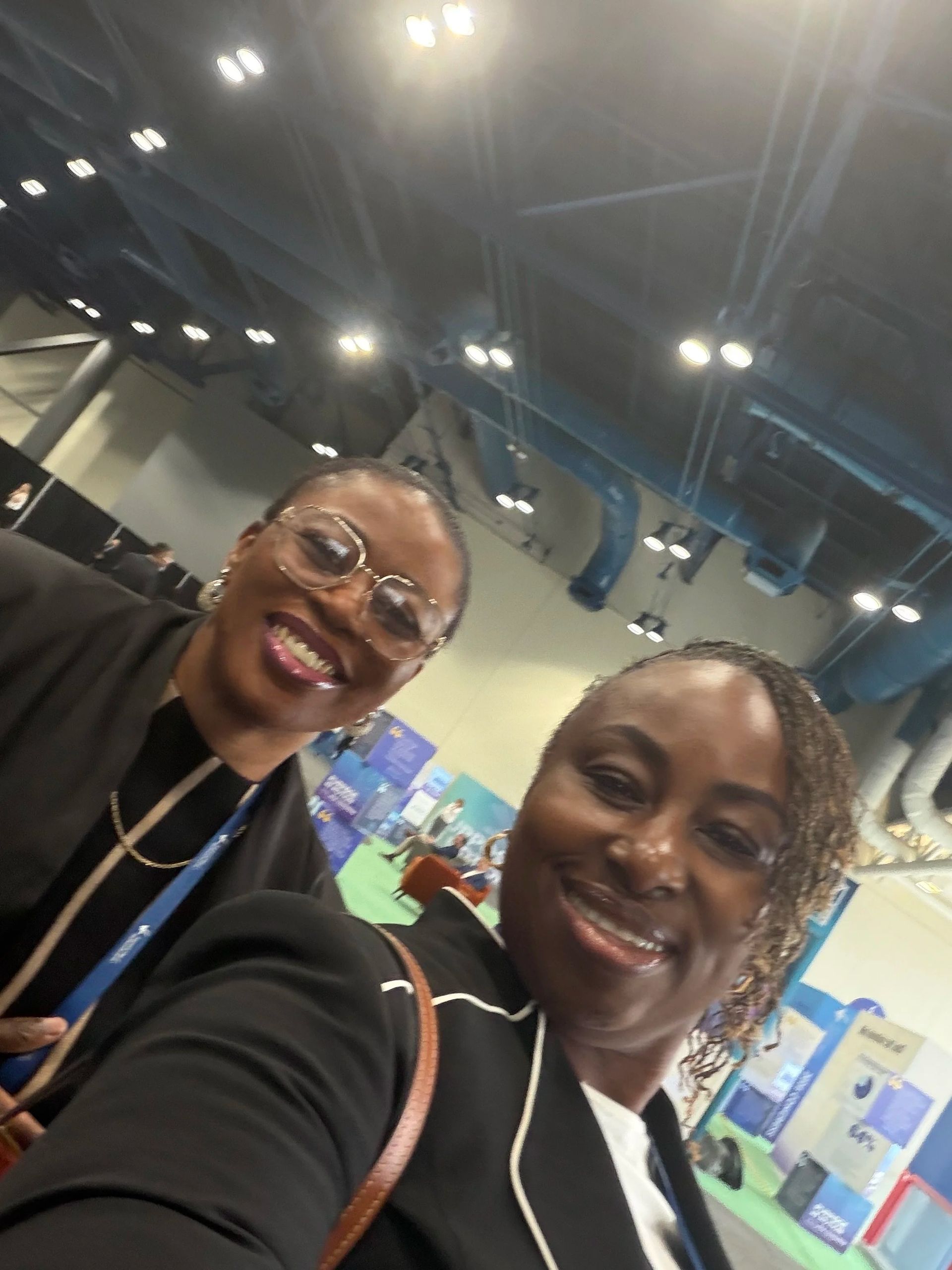 Two smiling people in a bright convention hall, taking a selfie with booths and lights in the background.