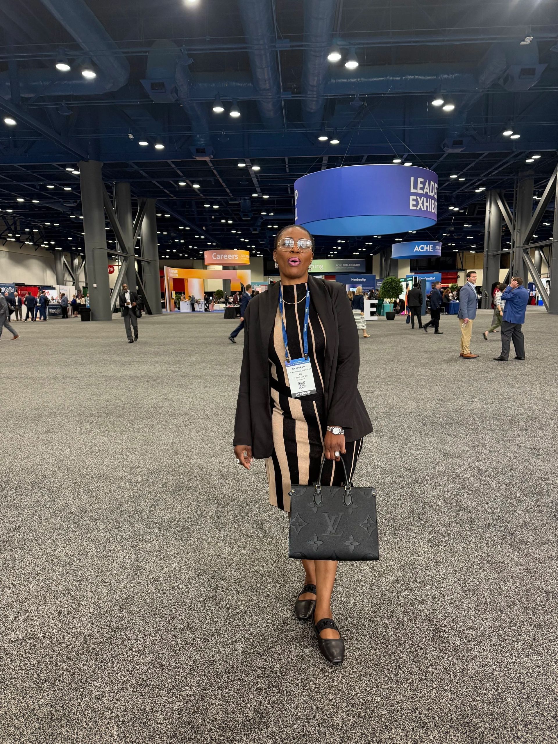 Person walking through a large convention hall, carrying a black handbag and wearing a black outfit.