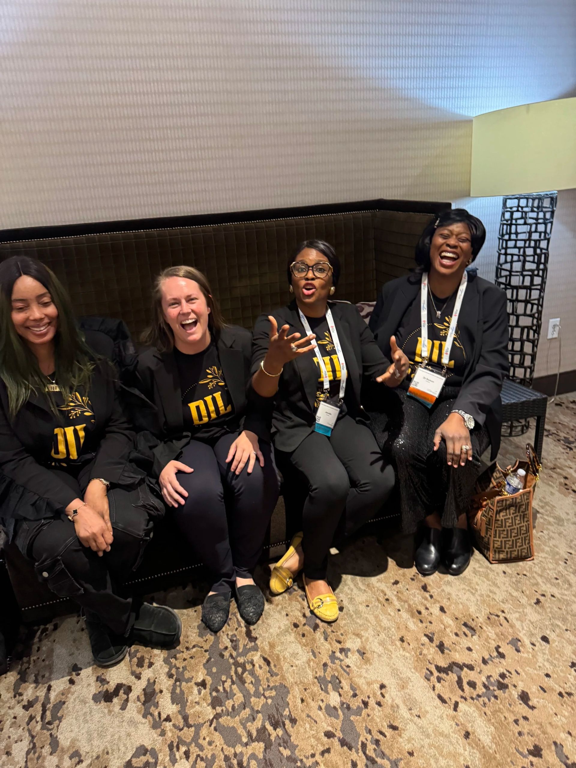 Four friends in black outfits sit smiling on a bench indoors, wearing lanyards and yellow accessories.