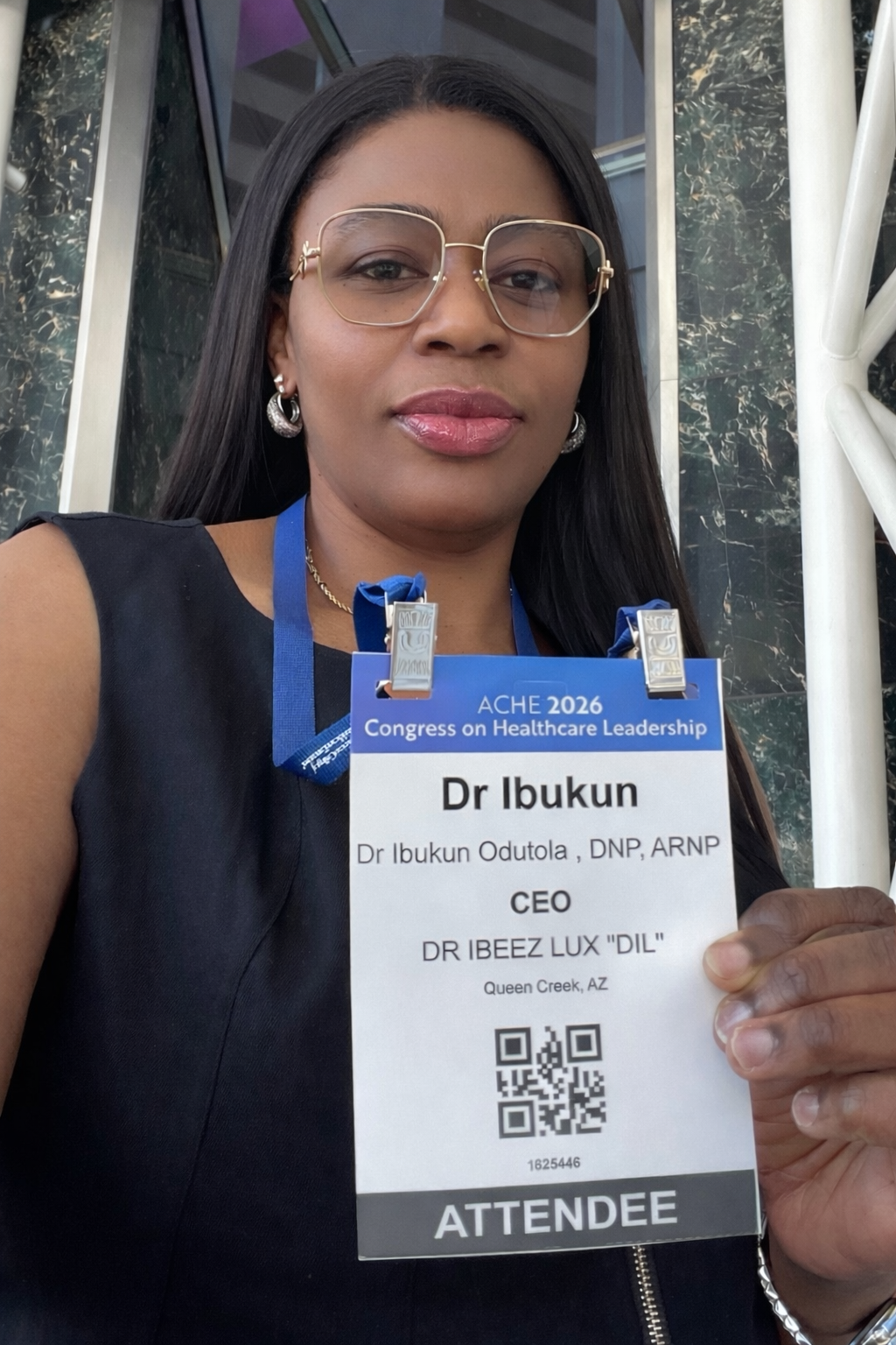 Woman holding an attendee badge, standing outdoors in a sleeveless navy top and glasses.