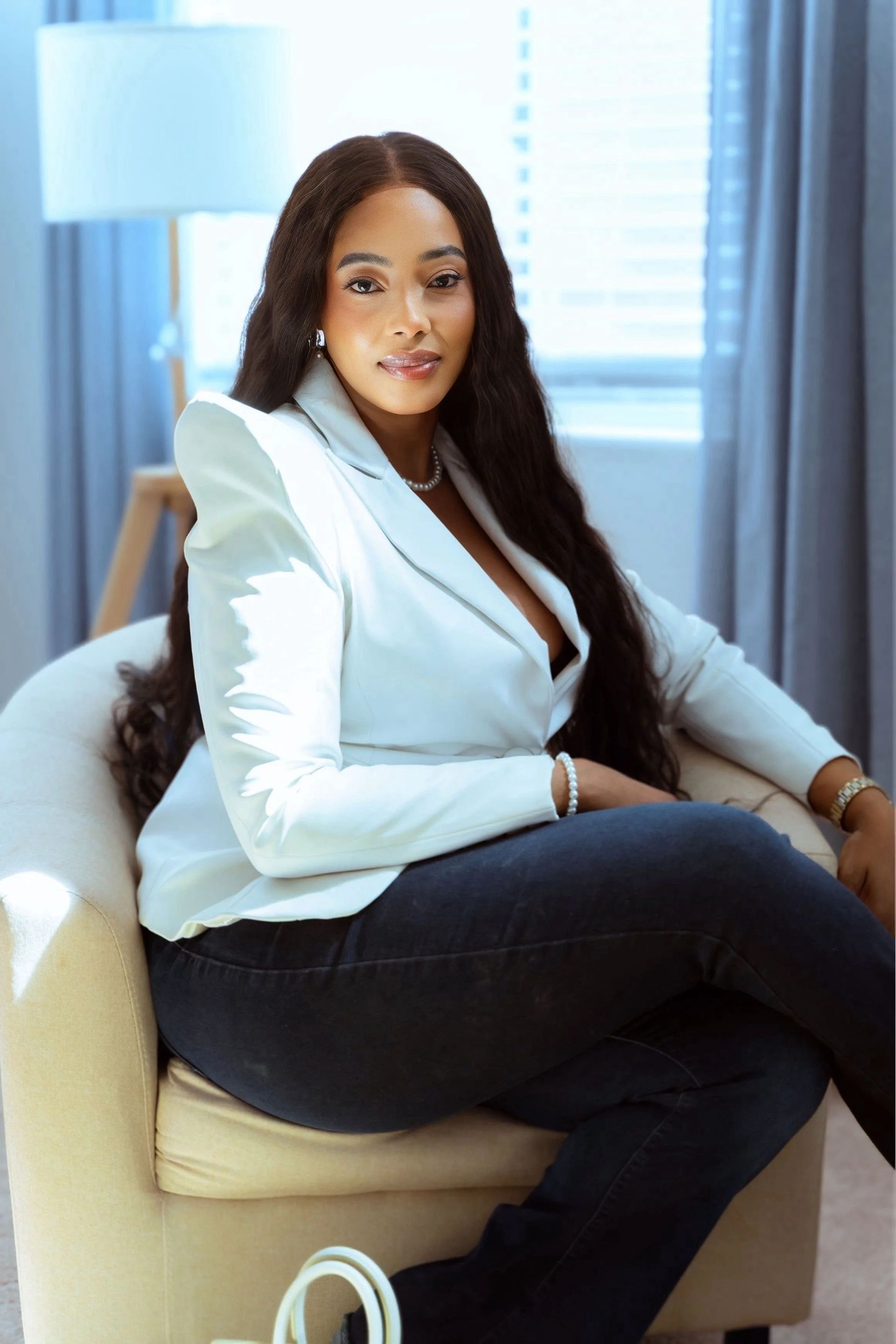Woman seated in a beige chair, wearing a light blue blazer and dark jeans, in a bright room by a window.