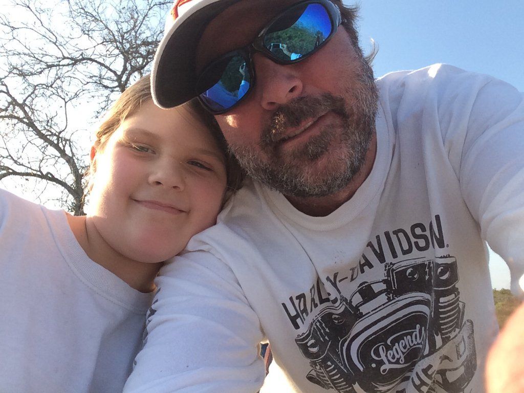 Close-up selfie of two people outdoors, smiling in sunlight, one wearing sunglasses and a cap.