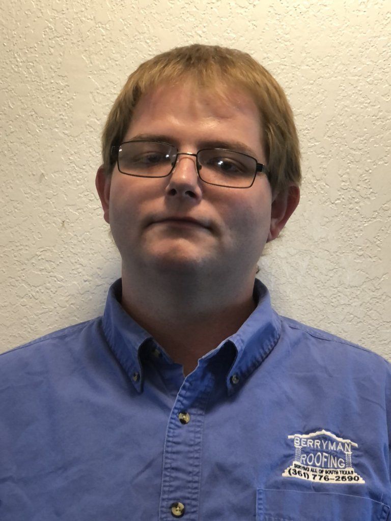 Person in a blue button-up shirt and glasses standing against a beige wall.