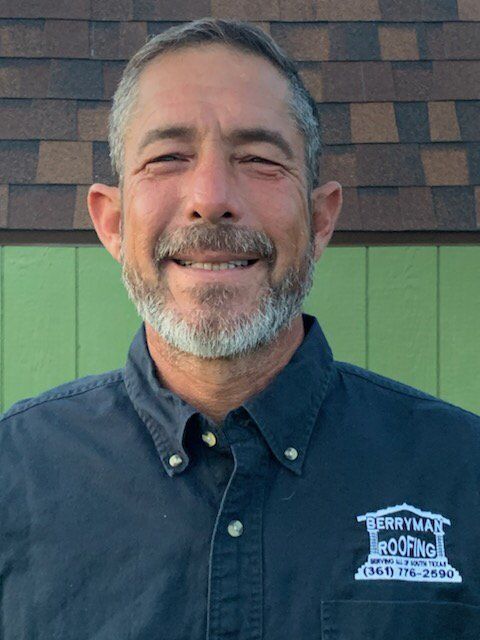 Smiling man in navy shirt standing outdoors in front of green wall and shingled roof.
