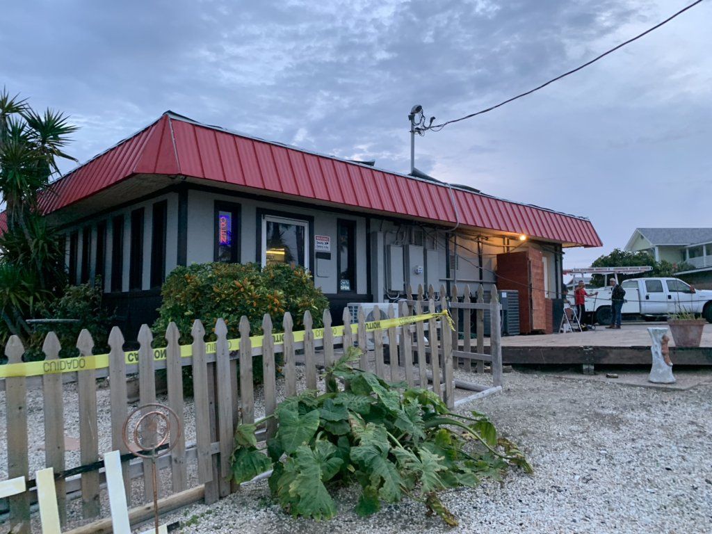 Small house with a red metal roof, picket fence, and gravel yard under a cloudy sky.