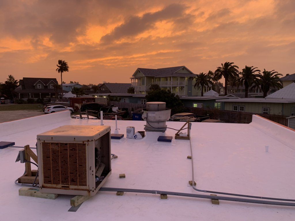 Rooftop at sunset with white equipment, palm trees, and houses silhouetted against an orange sky