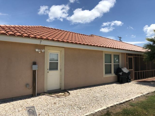Single-story beige house with red tile roof, white door, gravel path, and a barbecue grill under a blue sky.