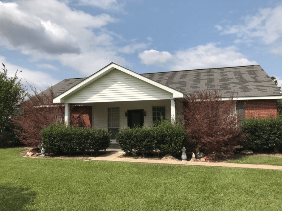 Single-story brick house with a front porch, white gable, and trimmed shrubs under a cloudy sky