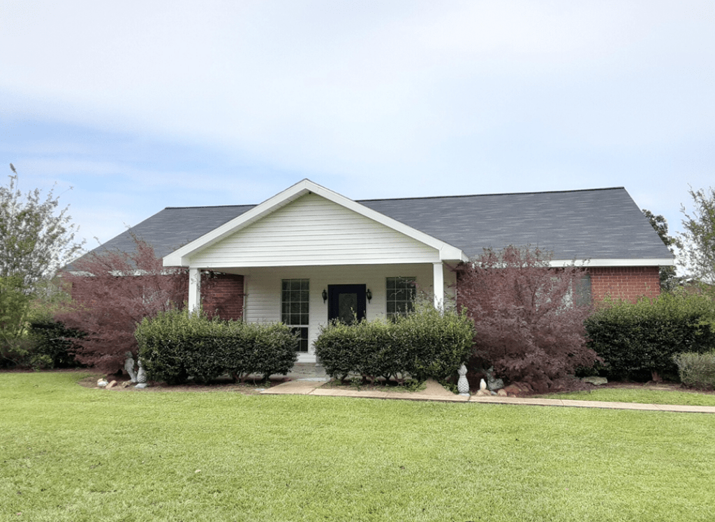 Single-story brick house with white porch, gray roof, and trimmed bushes in front of a green lawn