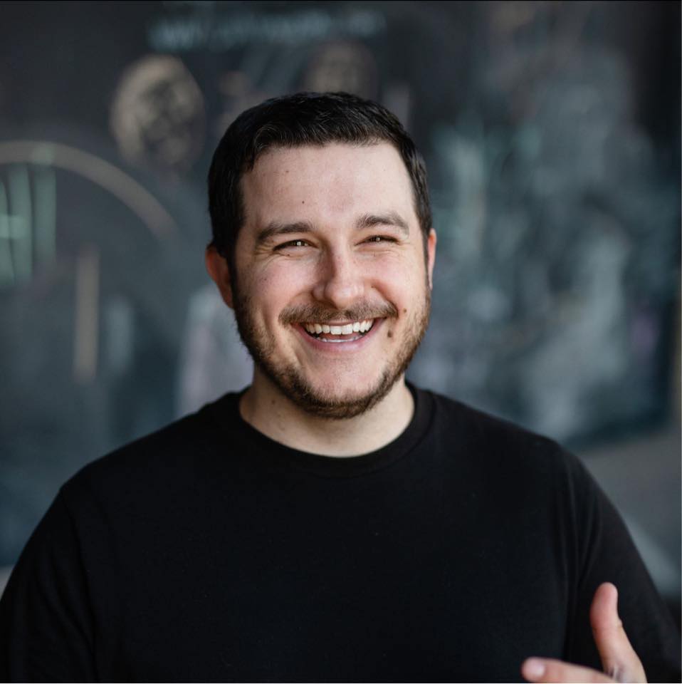 Josh Miller Man with brown hair and beard smiles broadly. He wears a black shirt, and a mural is in the background.