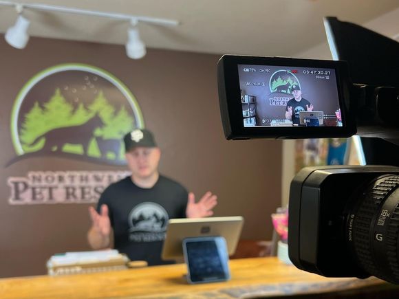 Man at desk in Northwest Pet Rescue studio, gesturing. Camera in foreground, logo on wall.