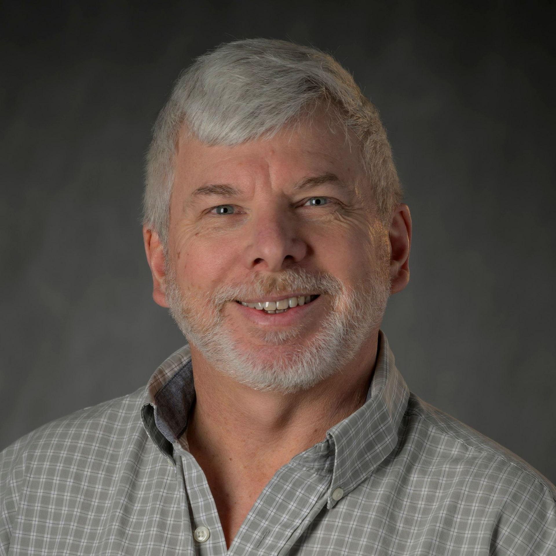 Travis Bertram Man with goatee, black cap, and black polo shirt, standing in front of a brick wall.
