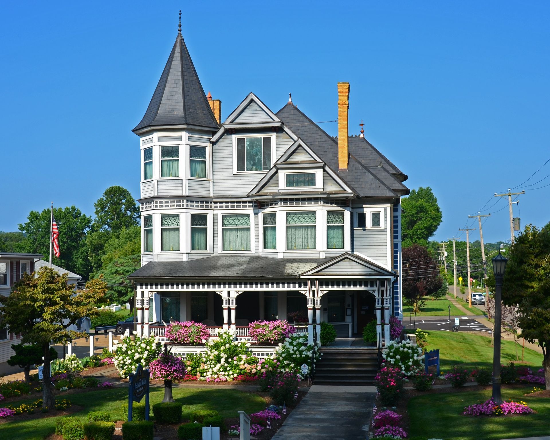 Victorian house with gray and white paint, a black turret, and a porch, set in a sunny, green landscape.