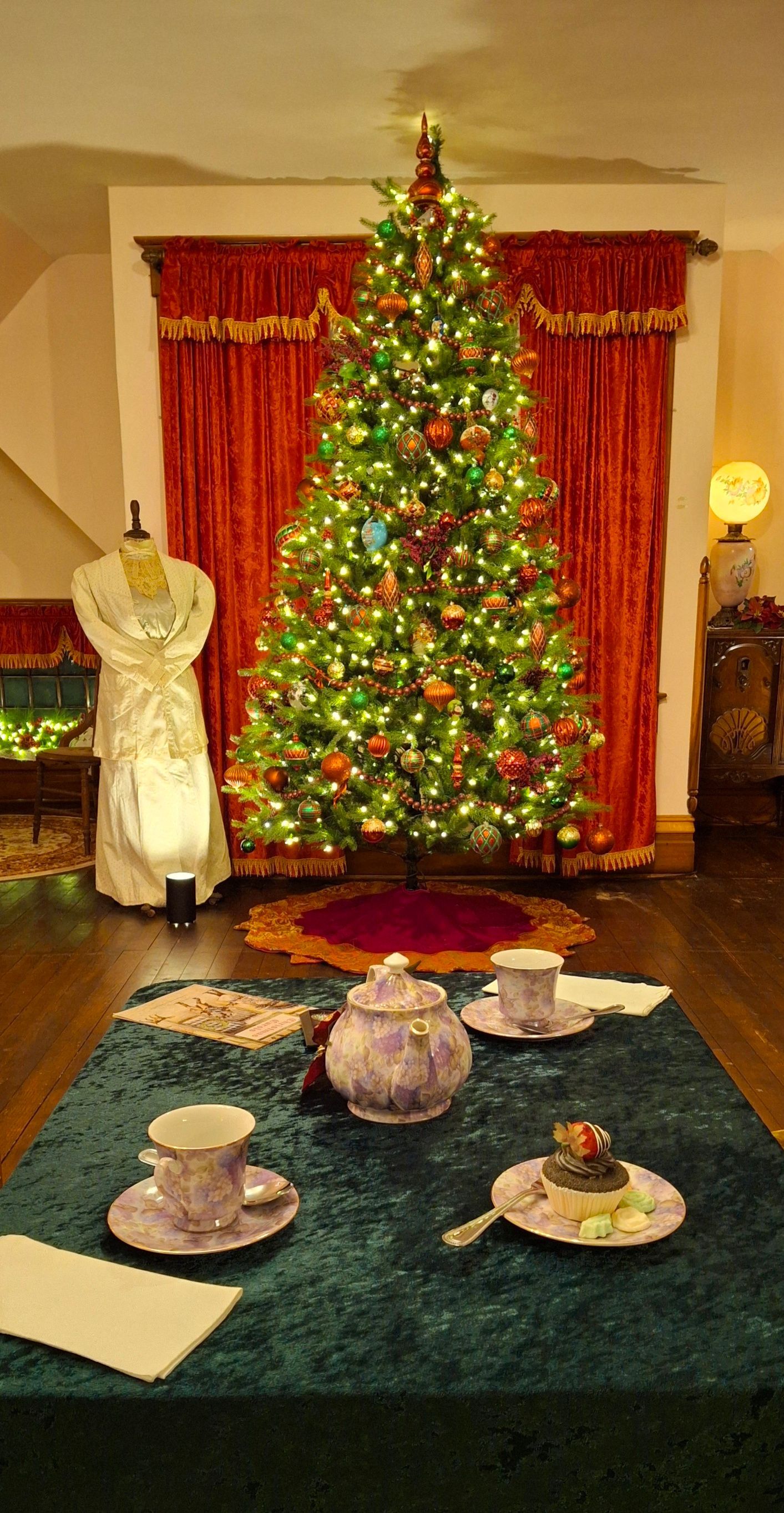 Christmas tree lit with ornaments, tea setting on a green tablecloth, elegant gown on a dress form, red curtains.