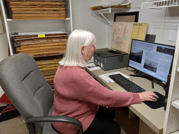 Woman seated at a desk, typing on a computer. A shelf with documents is behind her.