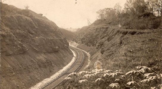 Railway tracks winding through a deep hillside cut, bordered by earthen walls and vegetation.