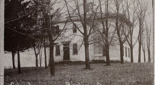 White two-story house with trees, in a grassy field.