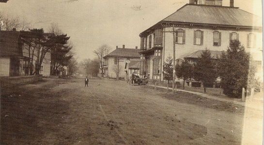 Dirt road in a small town. Buildings line the street, a man walks, a horse-drawn carriage sits.