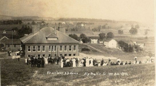 Historic school building with crowd outdoors; field in the foreground, distant houses and hills.