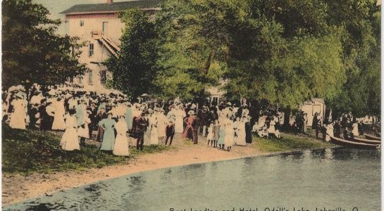 People gathered near a lake, some in white attire, near a building and trees.