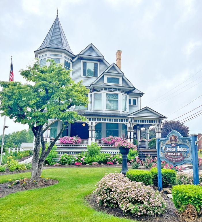 Victorian house with a turret, porch, and sign, surrounded by flowers and landscaping.