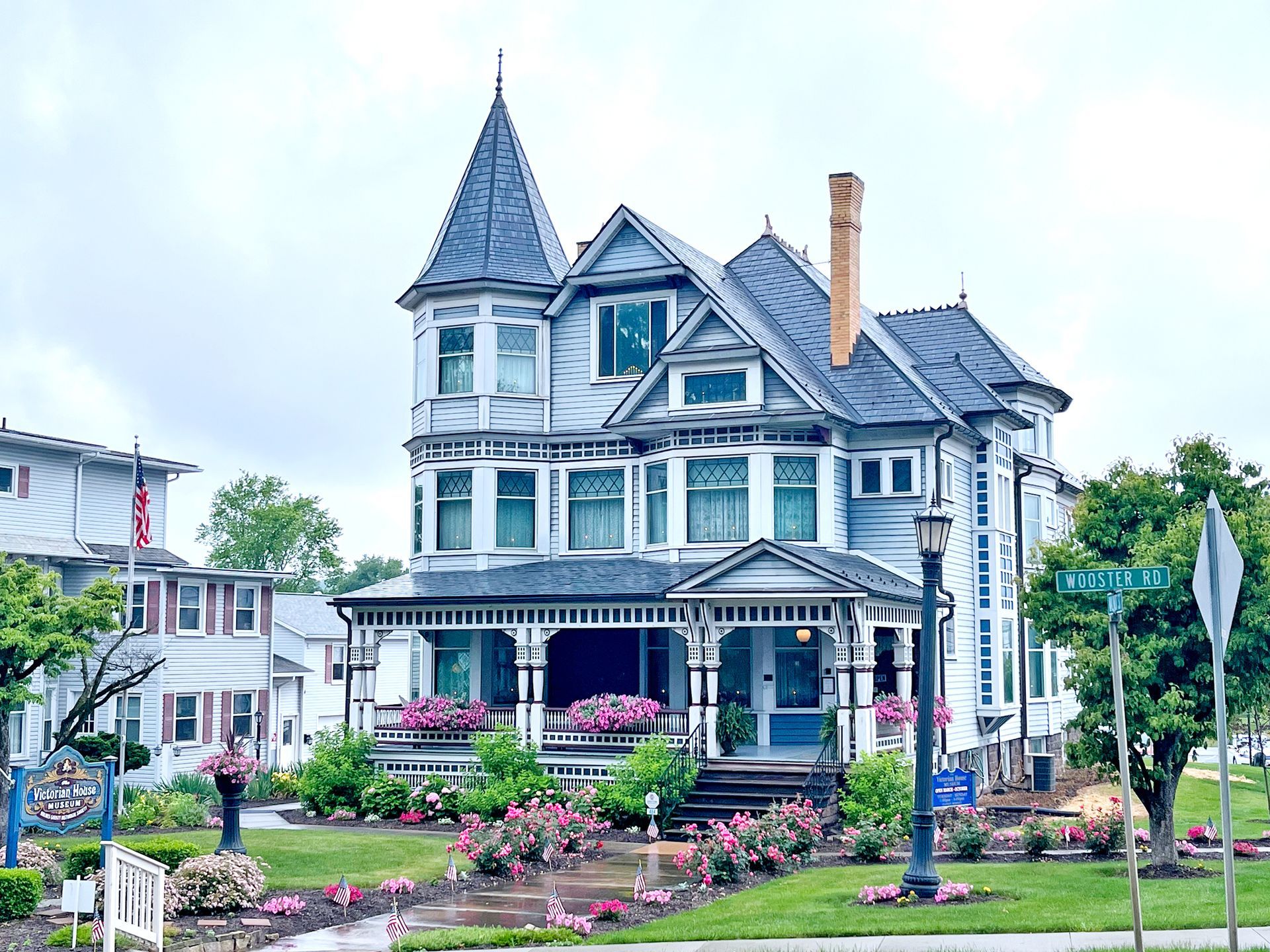 Victorian-style house, light blue exterior, turret, wraparound porch, and flowerbeds. Cloudy day.