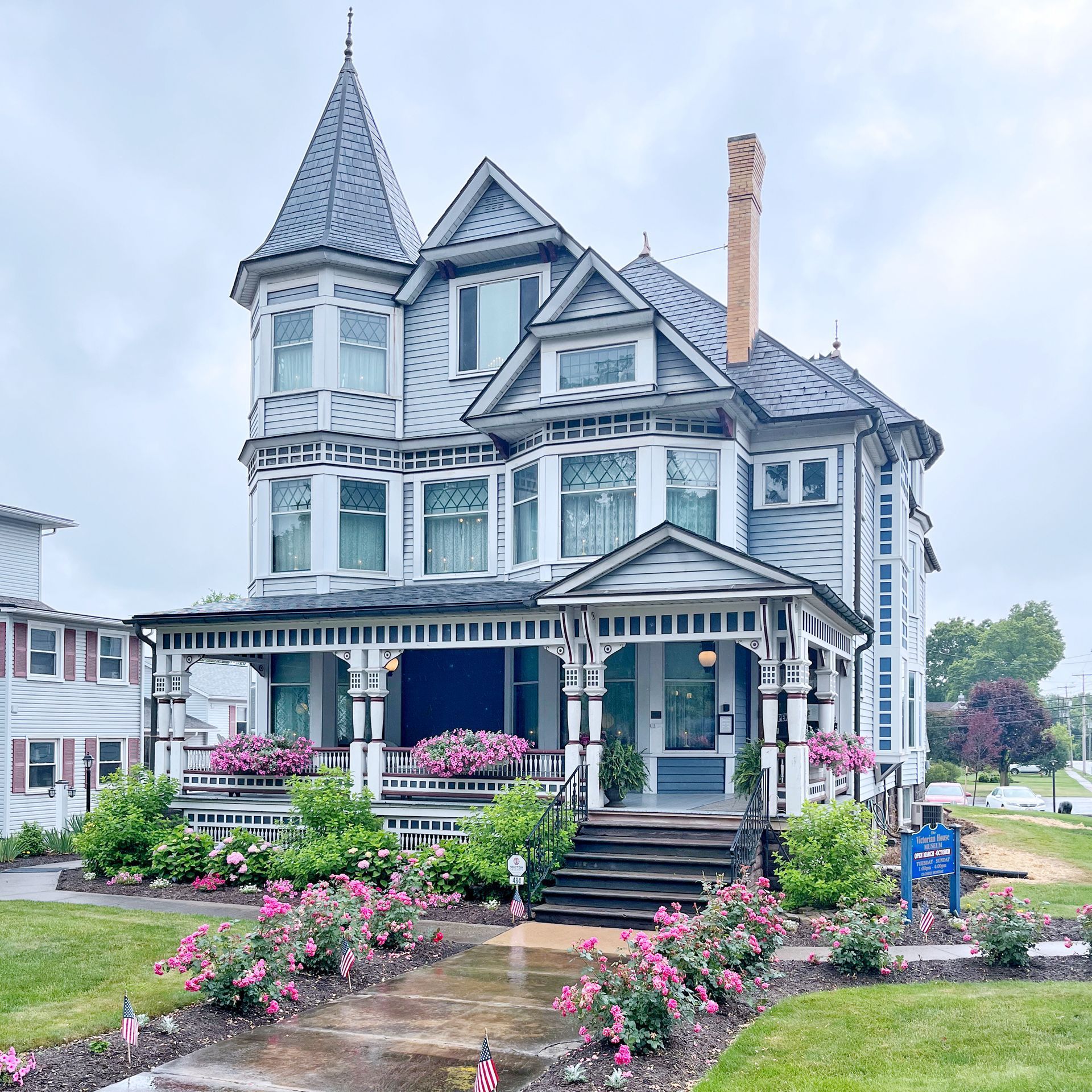 Ornate Victorian house with tower, blue and white paint, porch, flowers, and cloudy sky.