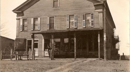 Old, two-story wooden building with porch and shutters. Dirt path leads to the front.