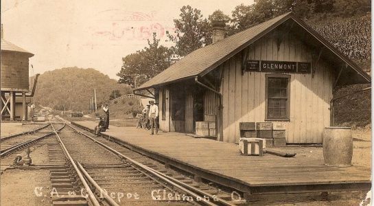 Vintage photograph of a train depot; two people on the platform, tracks, and a water tower in the background.