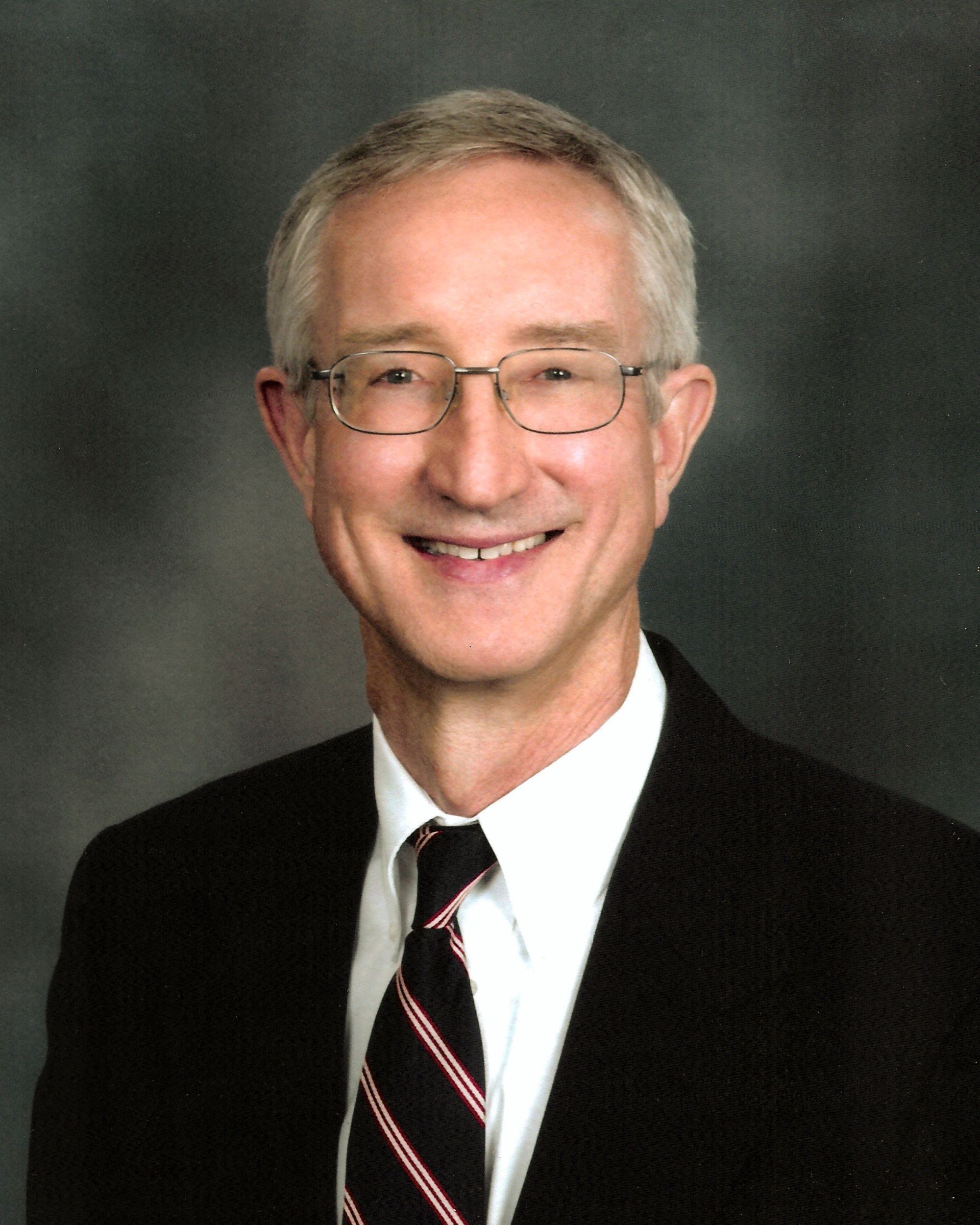 Man with glasses and tie smiles, wearing a suit jacket against a gray backdrop.