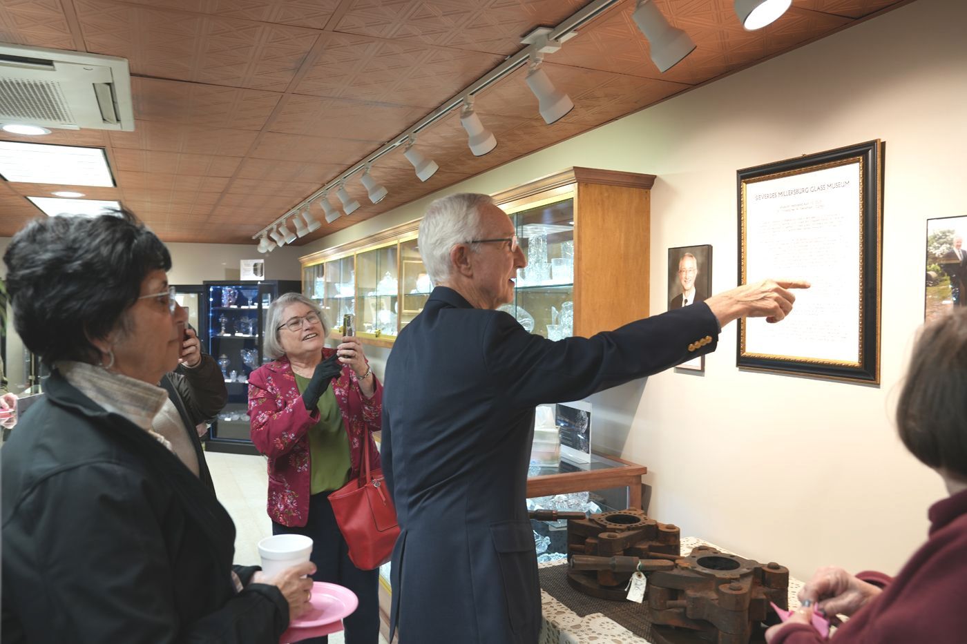 Man pointing at framed document, viewed by group inside a building with a display case and lighting.
