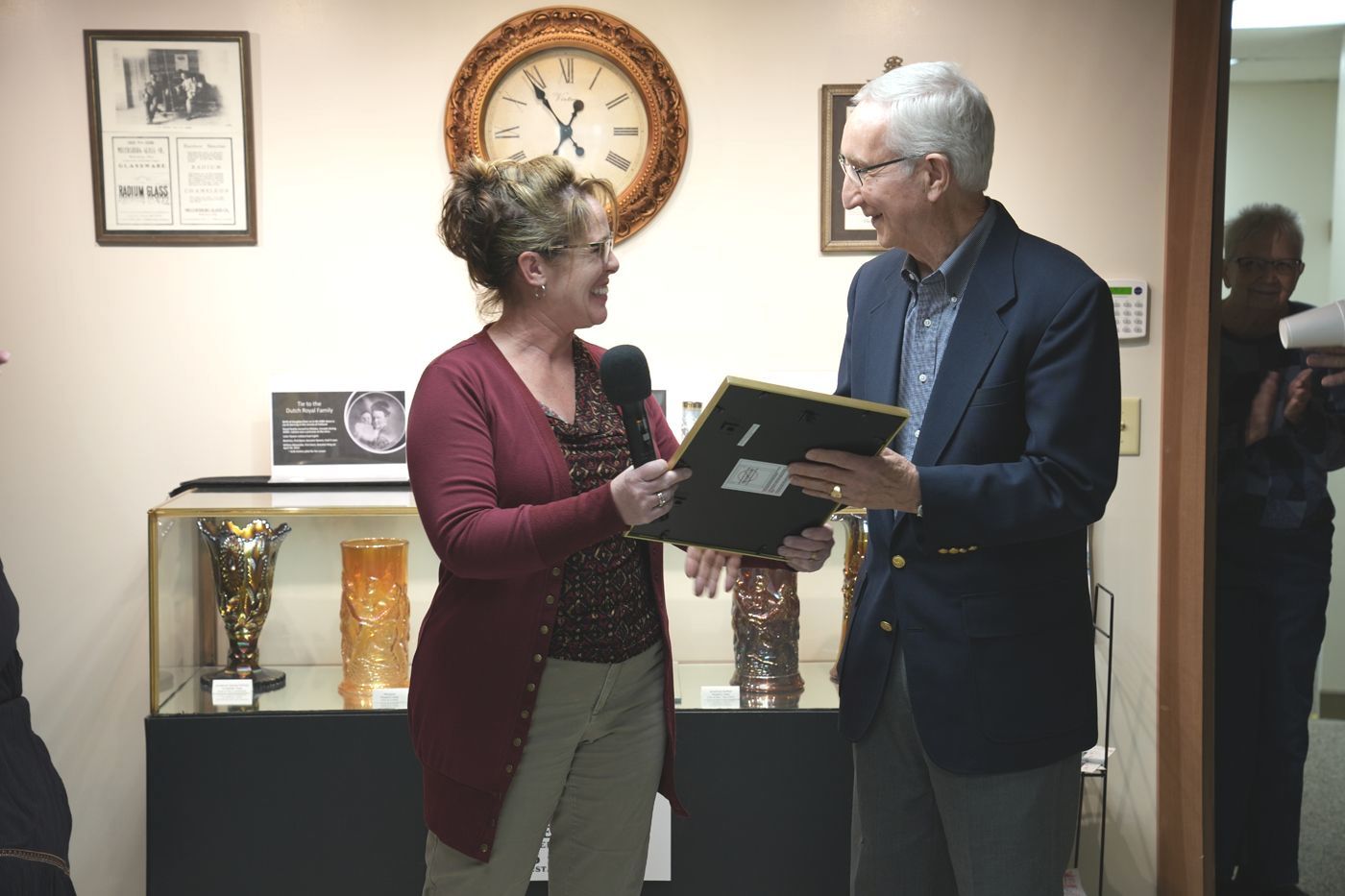 Woman handing a framed item to a man indoors, near a display case with vases and a clock on the wall.