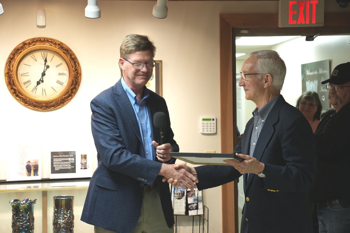 Two men shaking hands in a room, one holding a plaque, near a clock and exit sign.
