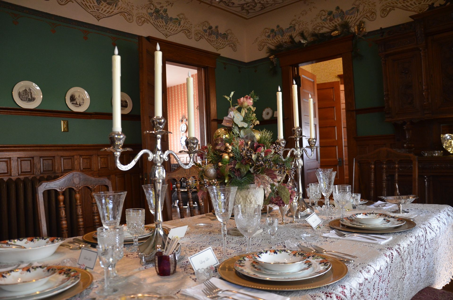 Formal dining room set for a meal. Green walls, wood trim, floral centerpiece, and lit candles.
