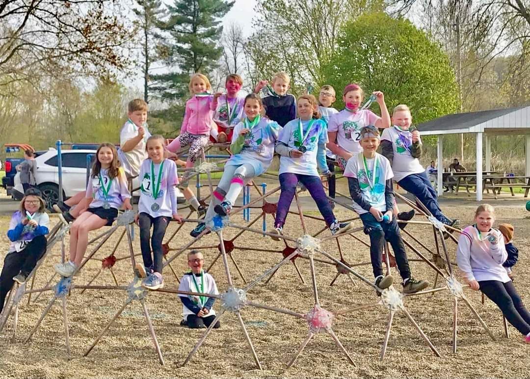 Group of kids on a metal playground structure; smiling and wearing tie-dye shirts, outdoors.
