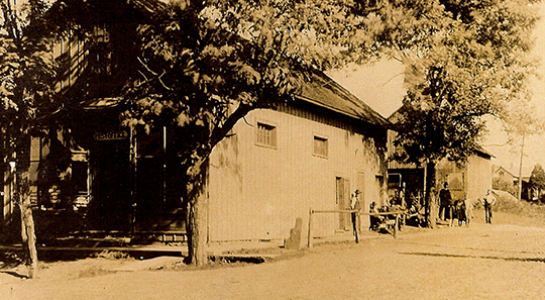 Old sepia photo of a wood building with a sign and people standing on a road. Tree in foreground.