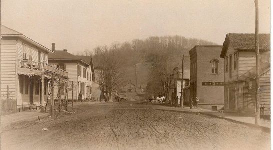 Old town street with wooden buildings on both sides, dirt road, hill in background.
