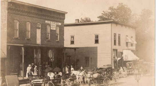 Historic photo of a town street scene with buildings, people, and horse-drawn carriages.