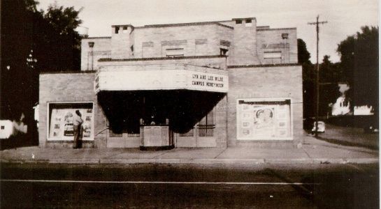 Brick building with awning over entrance, advertising signs on windows. A person stands near the doorway.