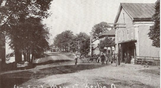 A vintage street scene with trees, buildings, and people walking on a dirt road.