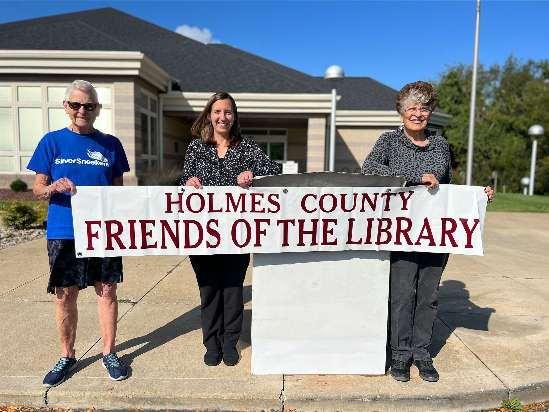 Three people holding a banner for Holmes County Friends of the Library in front of a library building.