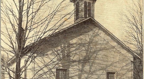 Old, weathered building with a small steeple, framed by bare tree branches; sepia tone.