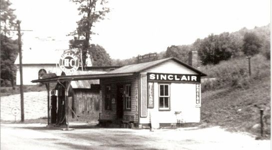 Vintage Sinclair gas station with visible pump and sign. Small building, rural setting, tree in the background.